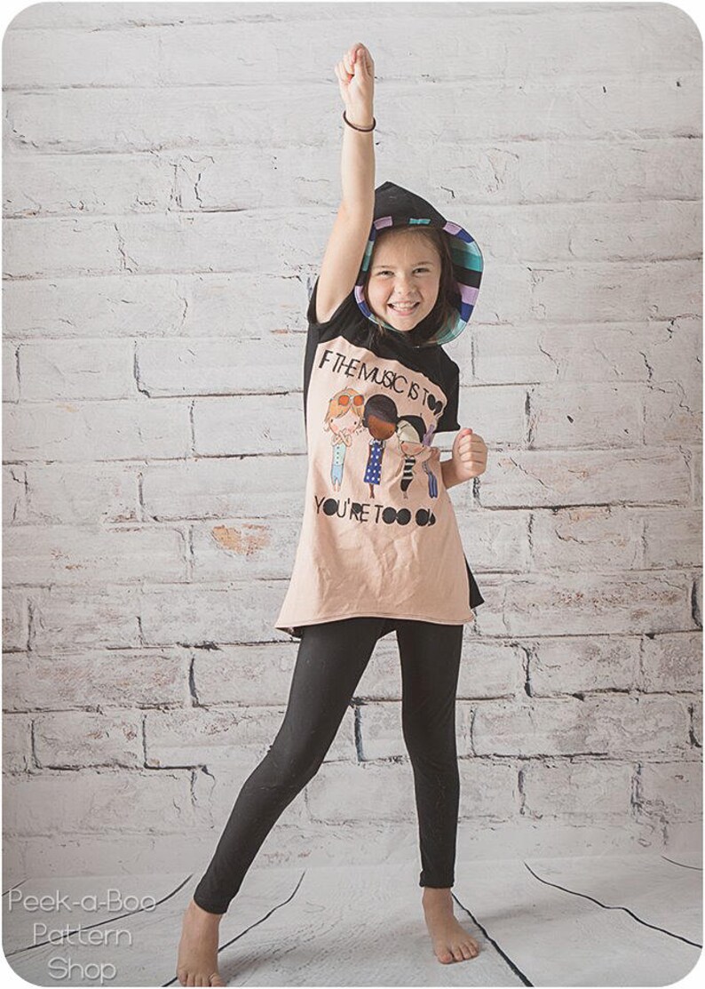 May include: A young person wearing a black and pink striped sun hat, a beige t-shirt with a graphic of four people and the text "If the music is too loud you're too old", and black leggings. The person is standing in front of a white brick wall.