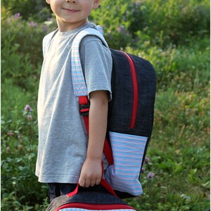 May include: A young boy wearing a gray t-shirt and a denim backpack with red accents. The backpack has a striped pocket and a matching striped lunch bag.