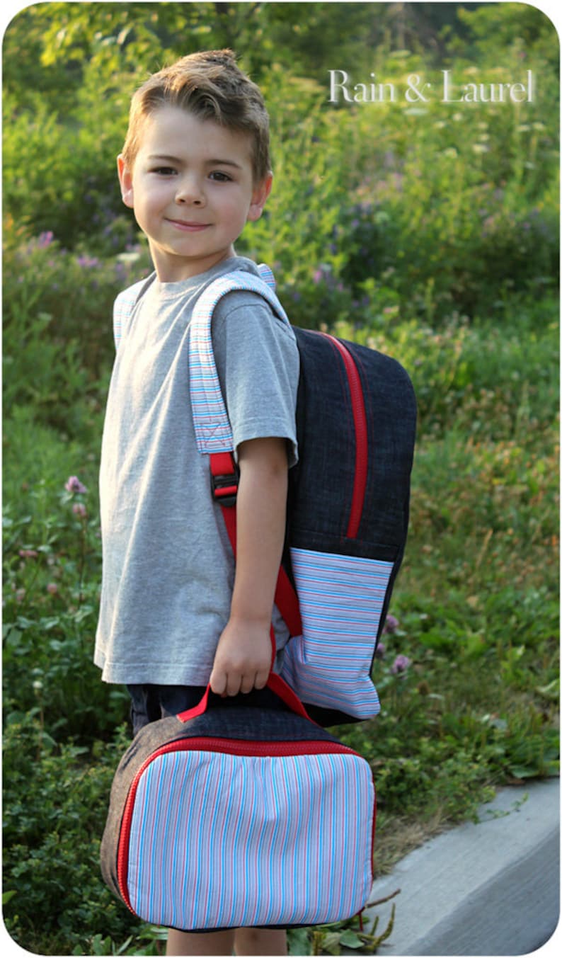 May include: A young boy wearing a gray t-shirt and a denim backpack with red accents. The backpack has a striped pocket and a matching striped lunch bag.