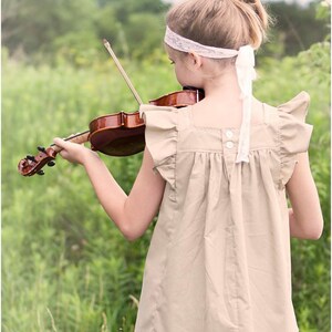 May include: A young person wearing a beige top with ruffled sleeves and a white headband plays a violin in a field of green grass.