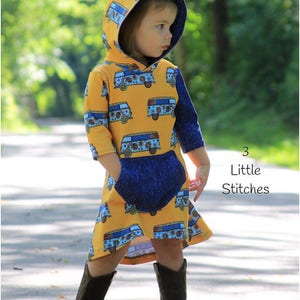 May include: A young girl wearing a yellow hooded dress with a colorful bus pattern and a blue pocket. The dress has long sleeves and a loose fit. The girl is wearing brown cowboy boots and is standing on a paved path.