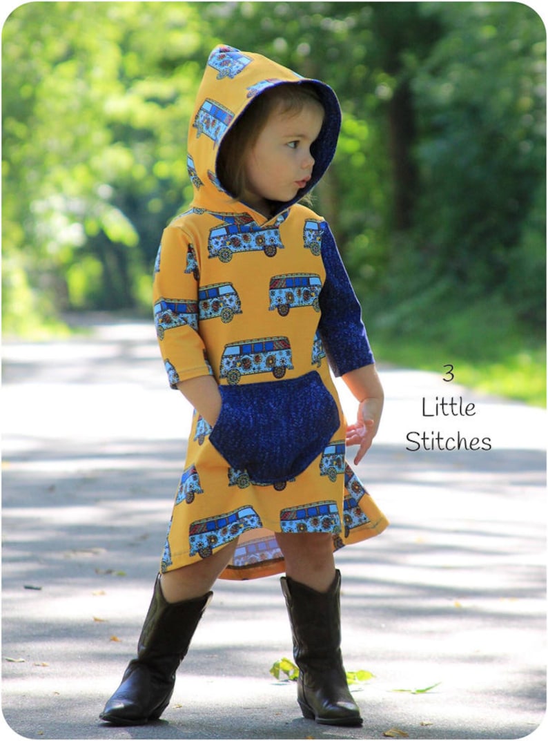 May include: A young girl wearing a yellow hooded dress with a colorful bus pattern and a blue pocket. The dress has long sleeves and a loose fit. The girl is wearing brown cowboy boots and is standing on a paved path.