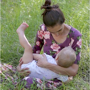 May include: A woman wearing a purple floral dress is sitting in a grassy field and holding a baby. The baby is wearing a white onesie with blue polka dots.