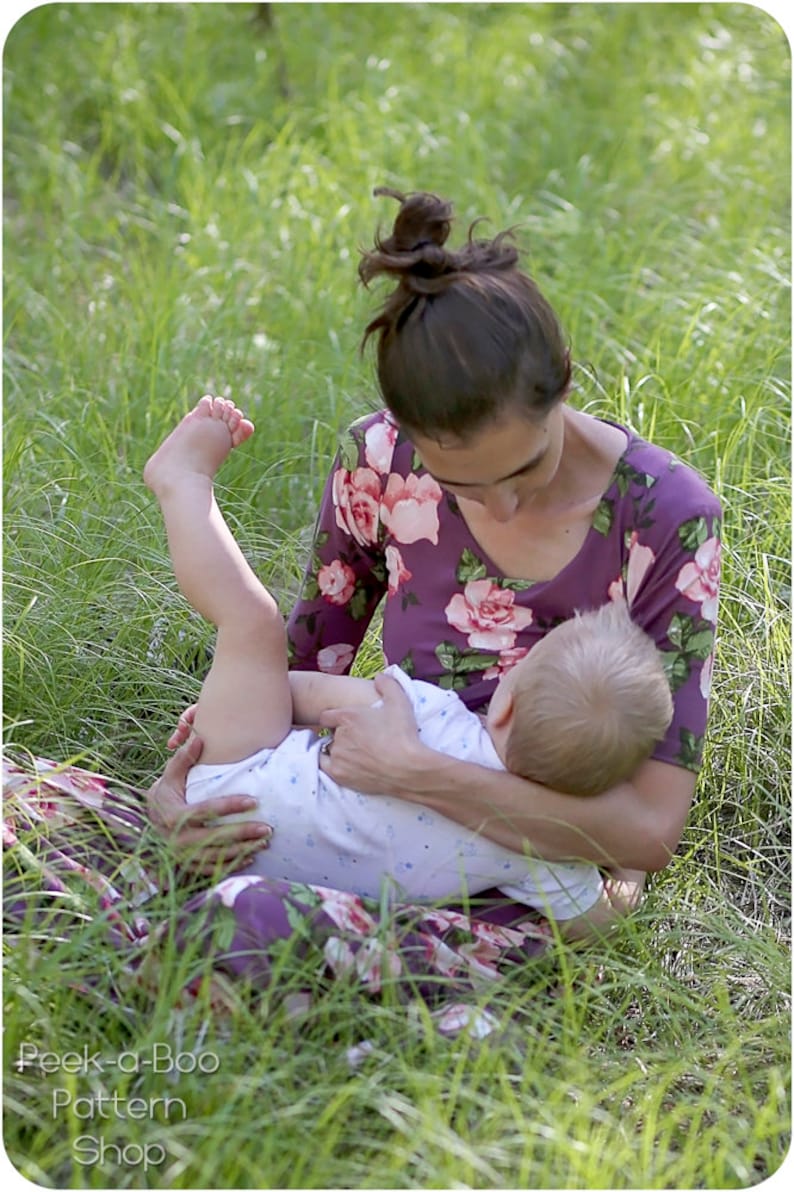May include: A woman wearing a purple floral dress is sitting in a grassy field and holding a baby. The baby is wearing a white onesie with blue polka dots.