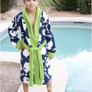 May include: A young person wearing a blue and green bathrobe with a white print of cartoon bears and the words "I love Bacon!" The robe has a green sash and a hood. The person is standing near a swimming pool and wearing blue and green slippers.