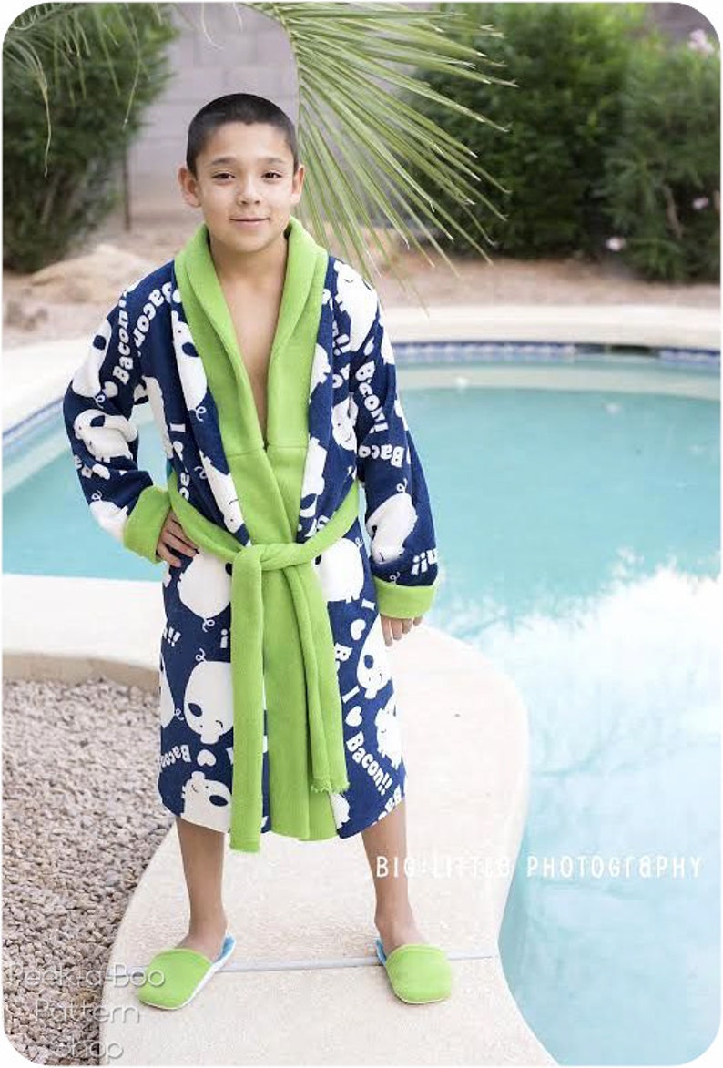 May include: A young person wearing a blue and green bathrobe with a white print of cartoon bears and the words "I love Bacon!" The robe has a green sash and a hood. The person is standing near a swimming pool and wearing blue and green slippers.