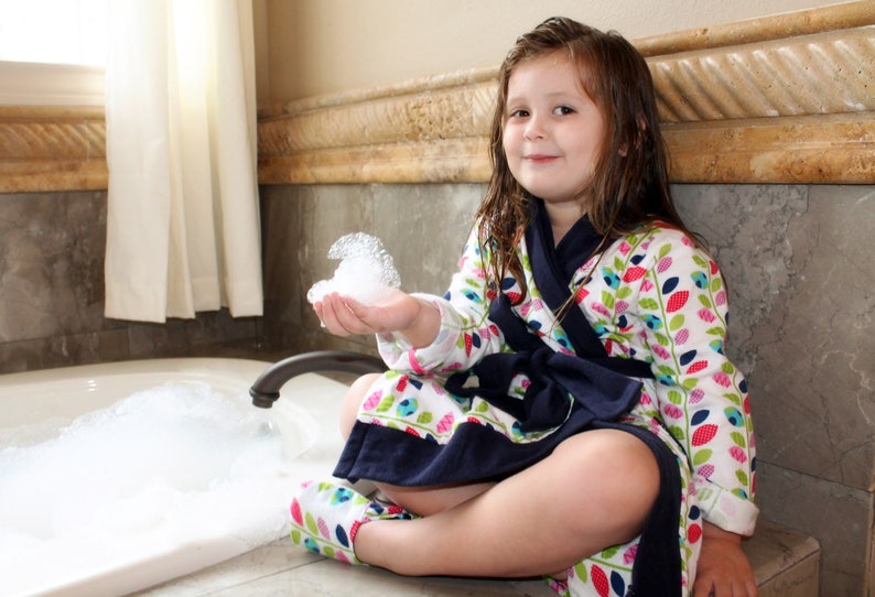May include: A young person wearing a white and blue floral robe with a navy blue sash sits on the edge of a bathtub filled with bubbles. The person is holding a handful of bubbles in their hand.