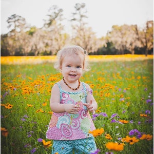May include: A young girl wearing a colorful patterned dress and shorts stands in a field of yellow and purple flowers. She is holding a small flower in her hand and smiling. The text "Simply Organic" is visible in the lower right corner of the image.