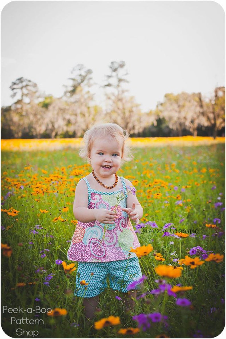 May include: A young girl wearing a colorful patterned dress and shorts stands in a field of yellow and purple flowers. She is holding a small flower in her hand and smiling. The text "Simply Organic" is visible in the lower right corner of the image.