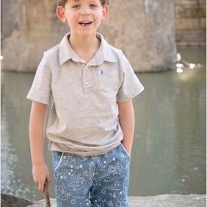 May include: A young boy wearing a grey polo shirt and blue shorts with a white map pattern. He is holding a stick in his right hand and smiling at the camera.