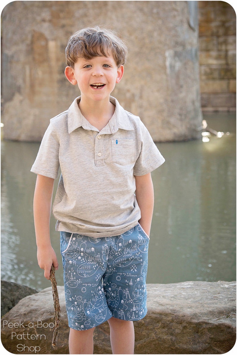 May include: A young boy wearing a grey polo shirt and blue shorts with a white map pattern. He is holding a stick in his right hand and smiling at the camera.