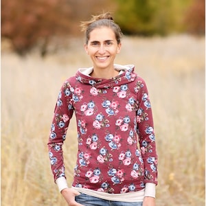 May include: A woman wearing a long-sleeved, burgundy floral print hooded sweatshirt with a white undershirt and blue jeans. She is standing in a field of tall grass.