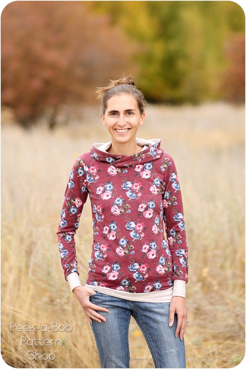 May include: A woman wearing a long-sleeved, burgundy floral print hooded sweatshirt with a white undershirt and blue jeans. She is standing in a field of tall grass.