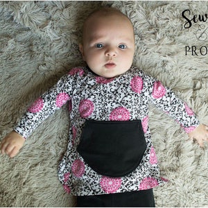 May include: A baby wearing a black and white long-sleeved shirt with pink floral accents and a black pocket. The shirt has a damask pattern. The baby is lying on a fluffy, light brown surface. The text "Sew & Tell PROJECT" is visible in the top right corner of the image.