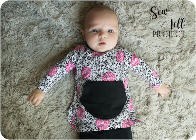 May include: A baby wearing a black and white long-sleeved shirt with pink floral accents and a black pocket. The shirt has a damask pattern. The baby is lying on a fluffy, light brown surface. The text "Sew & Tell PROJECT" is visible in the top right corner of the image.