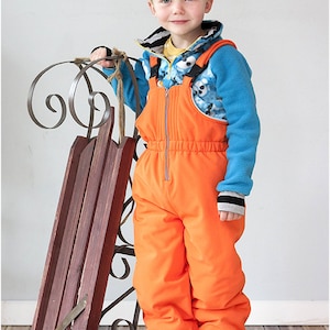 Puede incluir: Un niño pequeño con pantalones de nieve naranja, una chaqueta de vellón azul y una camisa con estampado blanco y negro. Está sosteniendo un trineo de madera con asas de metal.