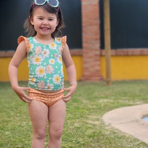 May include: A young girl wearing a green and white floral swimsuit with orange polka dot bottoms. She is smiling and wearing sunglasses. The girl is standing on a grassy lawn in front of a pool.