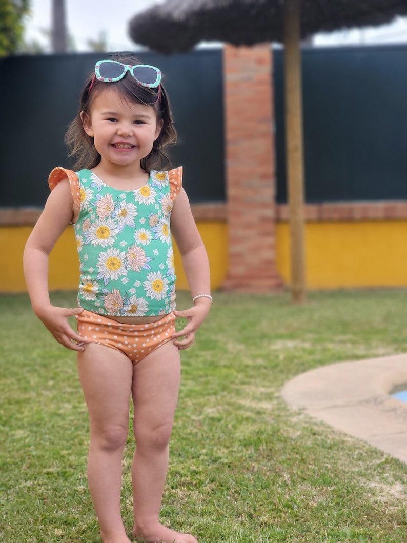 May include: A young girl wearing a green and white floral swimsuit with orange polka dot bottoms. She is smiling and wearing sunglasses. The girl is standing on a grassy lawn in front of a pool.