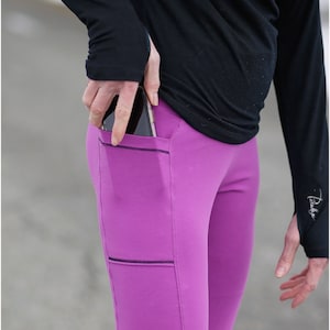 May include: Close-up of magenta leggings with black stitching and pockets. A person wearing a black long-sleeve shirt is putting a smartphone into the pocket. The leggings are part of the Peek-a-Boo Pattern Shop.
