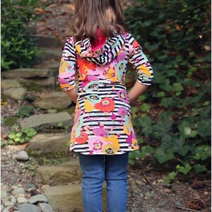 May include: A young person wearing a black and white striped hooded top with a floral print. The top has a pink hood and is worn over blue jeans. The person is standing on a path with rocks and green plants.
