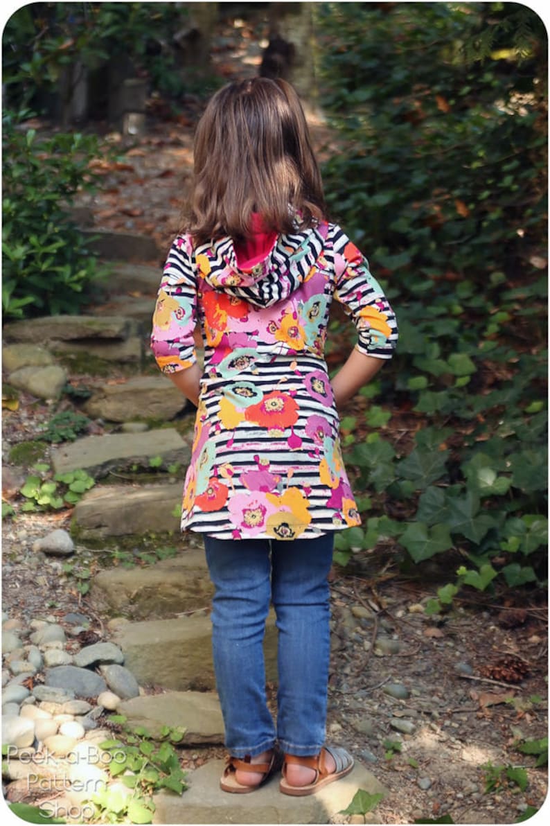 May include: A young person wearing a black and white striped hooded top with a floral print. The top has a pink hood and is worn over blue jeans. The person is standing on a path with rocks and green plants.