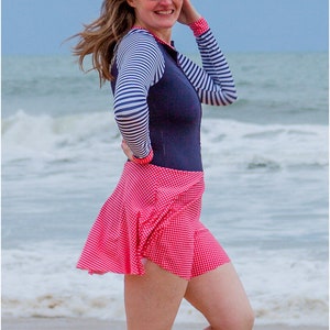 May include: A woman wearing a navy blue and white striped long-sleeved shirt with a red and white gingham skirt. She is standing on a beach with the ocean in the background.