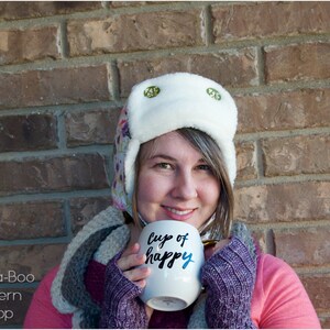 May include: A woman wearing a white and floral patterned winter hat with ear flaps and a pink sweater holds a white mug with the text "Cup of Happy" in black lettering.