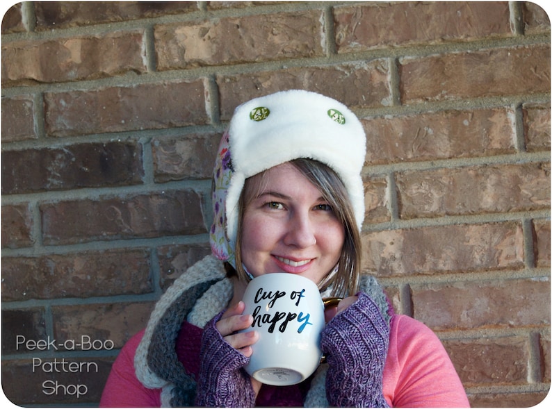 May include: A woman wearing a white and floral patterned winter hat with ear flaps and a pink sweater holds a white mug with the text "Cup of Happy" in black lettering.