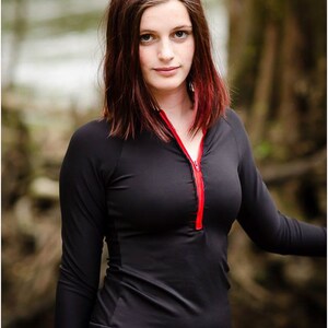 May include: A woman wearing a black long-sleeved shirt with a red zipper. The shirt has a fitted style and a high neckline.