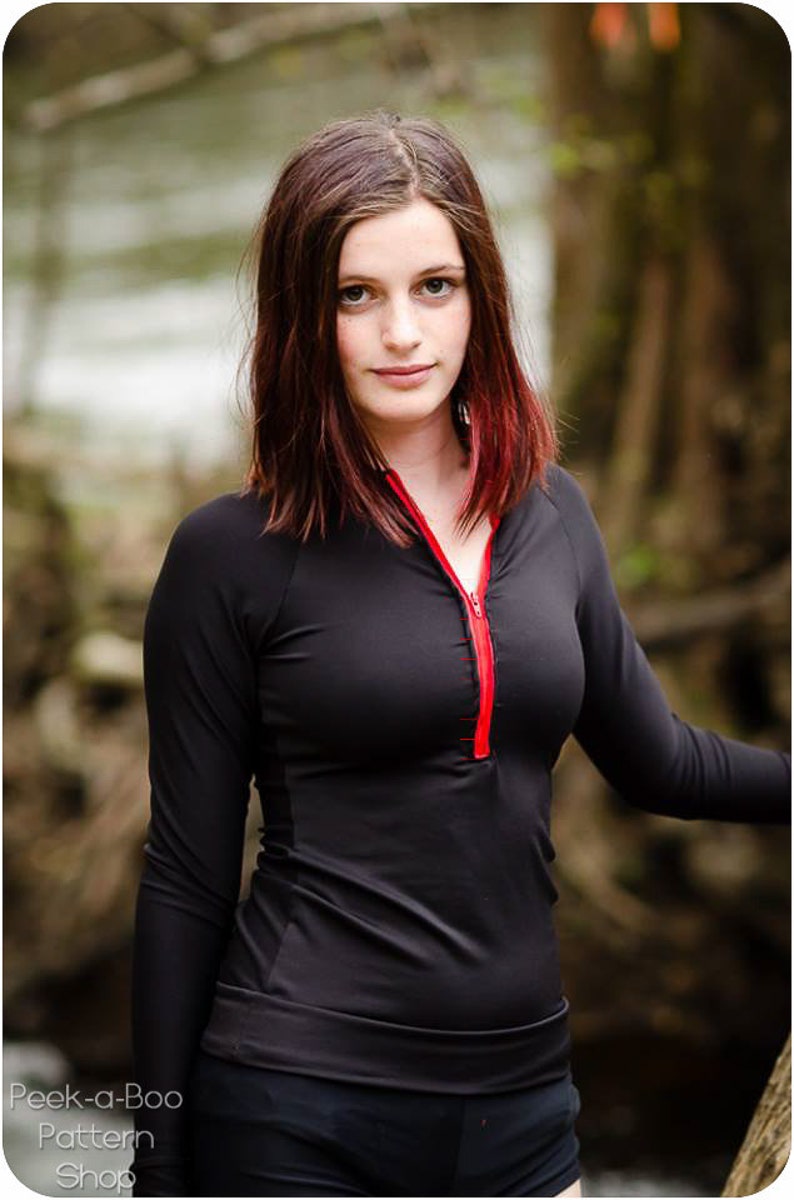 May include: A woman wearing a black long-sleeved shirt with a red zipper. The shirt has a fitted style and a high neckline.