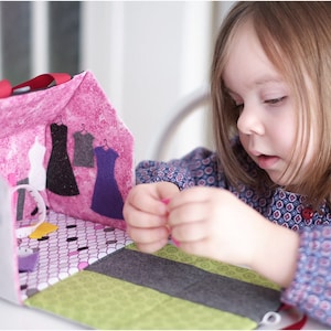 Peut inclure: Une jeune fille joue avec une maison de poup&eacute;e en feutre avec un int&eacute;rieur rose et un ext&eacute;rieur gris. La maison de poup&eacute;e a une petite fen&ecirc;tre et un sol en feutre avec un motif &agrave; carreaux noir et blanc. La maison de poup&eacute;e est ouverte et l'int&eacute;rieur montre un placard en feutre avec trois robes accroch&eacute;es &agrave; l'int&eacute;rieur.