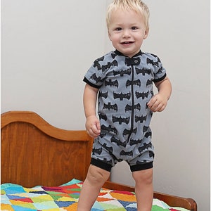 May include: A young child wearing a gray and black short-sleeved onesie with a bat pattern stands on a colorful patchwork quilt. The onesie has a zipper down the front.