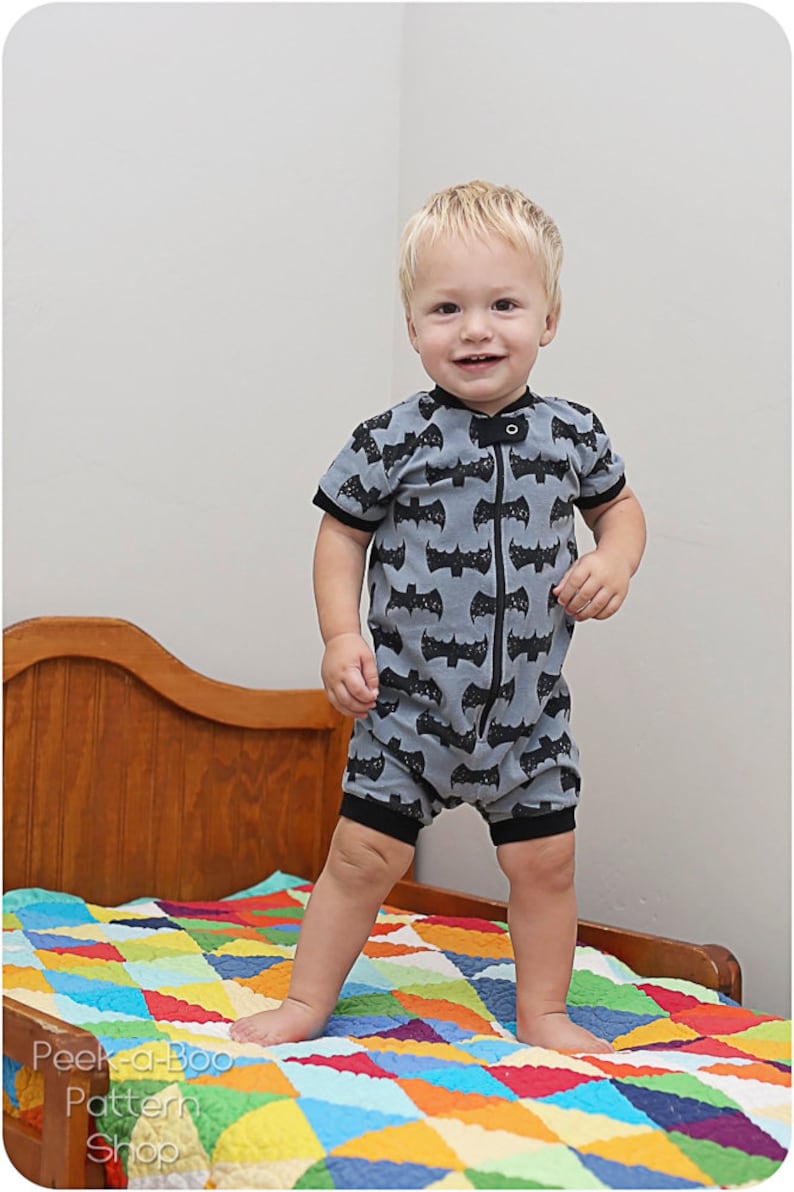 May include: A young child wearing a gray and black short-sleeved onesie with a bat pattern stands on a colorful patchwork quilt. The onesie has a zipper down the front.