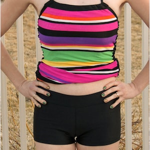 May include: A woman wearing a striped halter top and black swim shorts. The top has pink, white, orange, purple, green, and yellow stripes. The shorts are a solid black.