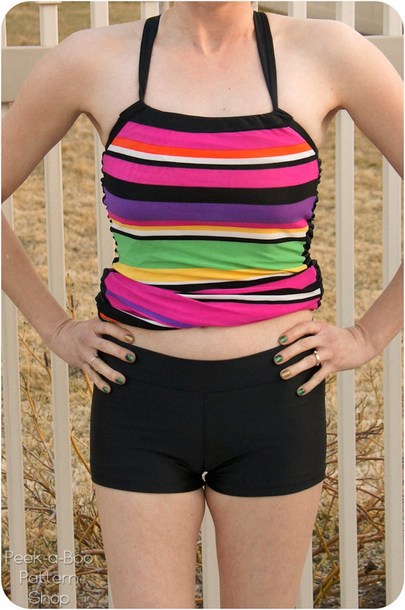 May include: A woman wearing a striped halter top and black swim shorts. The top has pink, white, orange, purple, green, and yellow stripes. The shorts are a solid black.