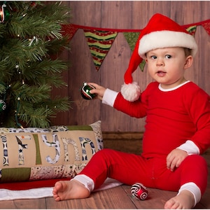 Puede incluir: Un ni&ntilde;o peque&ntilde;o con un traje de Pap&aacute; Noel rojo y un gorro de Pap&aacute; Noel se sienta frente a un &aacute;rbol de Navidad y sostiene un adorno verde. El &aacute;rbol est&aacute; decorado con luces y adornos. Una almohada con el nombre "Lilynn" est&aacute; en primer plano.