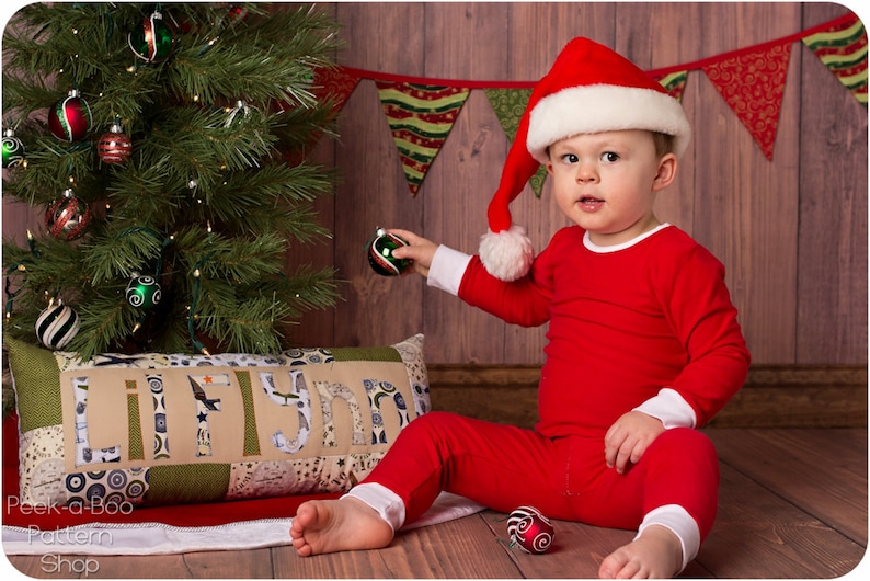 Puede incluir: Un ni&ntilde;o peque&ntilde;o con un traje de Pap&aacute; Noel rojo y un gorro de Pap&aacute; Noel se sienta frente a un &aacute;rbol de Navidad y sostiene un adorno verde. El &aacute;rbol est&aacute; decorado con luces y adornos. Una almohada con el nombre "Lilynn" est&aacute; en primer plano.