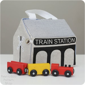 Peut inclure: Une gare en tissu ray&eacute;e bleu et blanc avec une pancarte noire indiquant "Gare".  Il y a trois trains jouets devant la gare : une locomotive rouge, un wagon jaune et un wagon rouge.