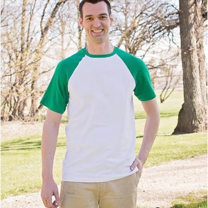 May include: A man wearing a white and green baseball tee shirt with khaki shorts. He is standing in a grassy area with trees in the background. The text "Sew & Tell Project" is visible in the lower right corner of the image.