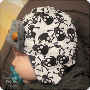 May include: A baby wearing a white and black skull print hat with a gray brim. The hat has a wooden button on the side.