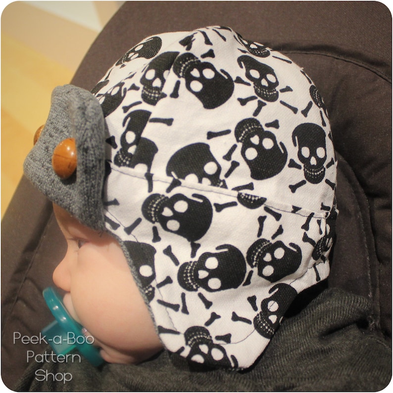 May include: A baby wearing a white and black skull print hat with a gray brim. The hat has a wooden button on the side.