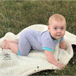 May include: A baby in a gray and turquoise onesie is crawling on a white fluffy blanket. The baby is looking directly at the camera with a curious expression. The blanket is on a bed of green grass.