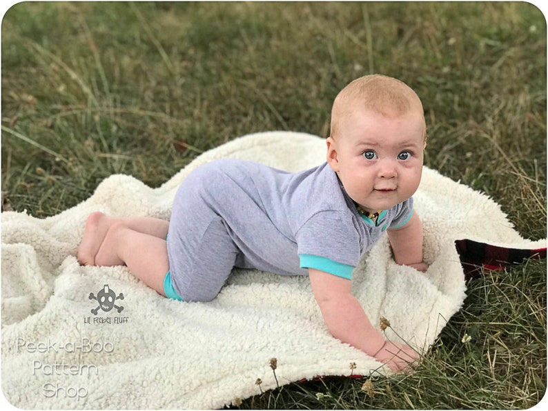 May include: A baby in a gray and turquoise onesie is crawling on a white fluffy blanket. The baby is looking directly at the camera with a curious expression. The blanket is on a bed of green grass.
