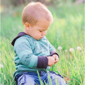 May include: A young child wearing a blue and green striped hoodie with black sleeves sits in a field of green grass, looking down at a blade of grass in their hands.