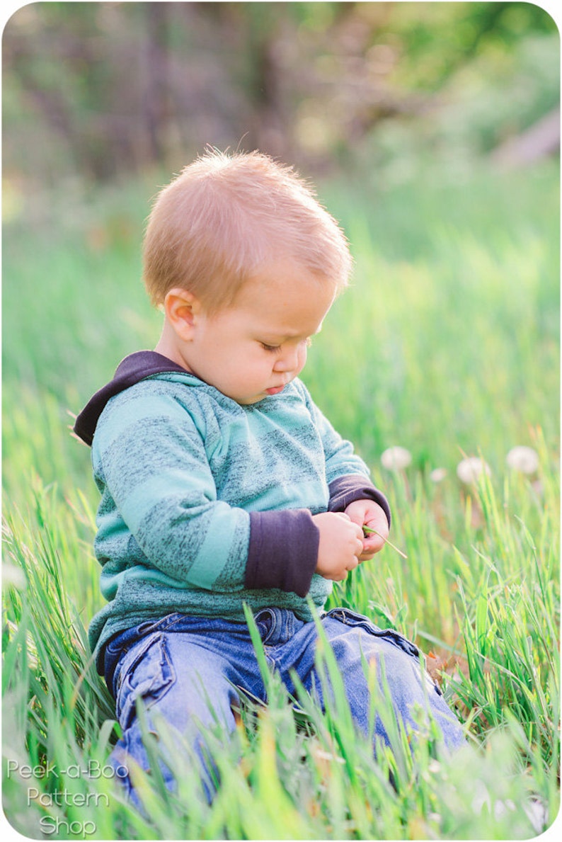 May include: A young child wearing a blue and green striped hoodie with black sleeves sits in a field of green grass, looking down at a blade of grass in their hands.