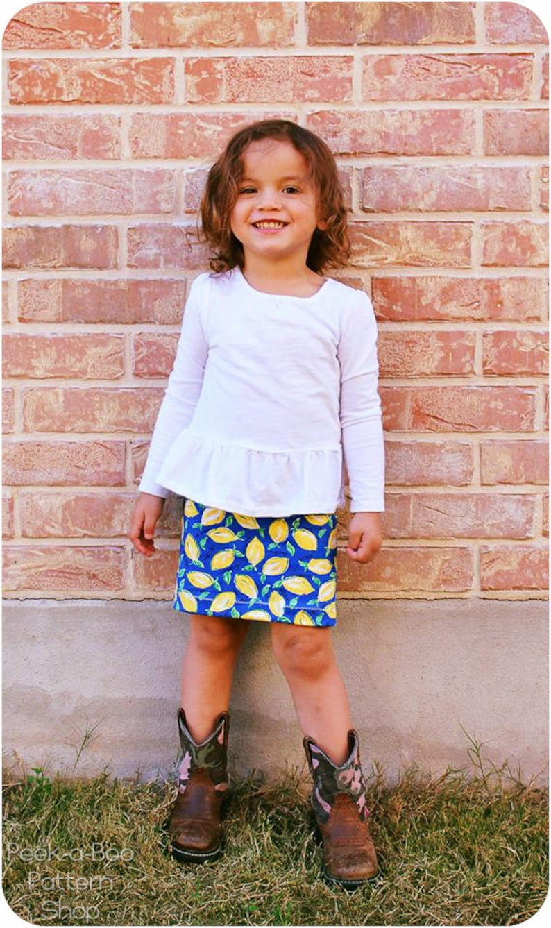 Puede incluir: Una ni&ntilde;a peque&ntilde;a con una camisa blanca de manga larga con un dobladillo con volantes y una falda con estampado de limones azules y amarillos. Est&aacute; de pie frente a una pared de ladrillo y lleva botas de vaquero marrones con detalles rosas y verdes.