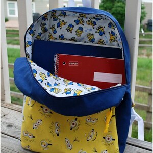 May include: Blue backpack with a yellow fabric lining featuring a repeating pattern of Minions. The backpack is open and has a red notebook and a blue notebook inside.
