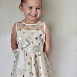 May include: A young girl wearing a white dress with gold polka dots and a white satin sash. She is smiling and has a white bow in her hair.