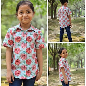 Puede incluir: Una persona joven lleva una camisa de manga corta de color azul claro con botones y un estampado floral rosa y rojo. La camisa tiene cuello y cierre de botones. La persona lleva pantalones vaqueros azules.