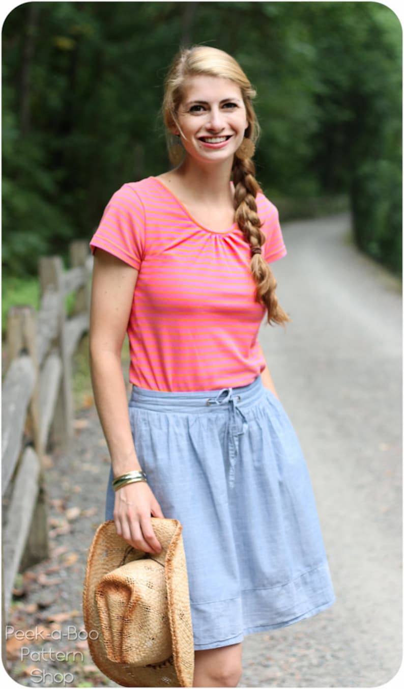 May include: A woman wearing a pink and orange striped short-sleeved shirt and a light blue skirt. She is holding a straw hat in her right hand.